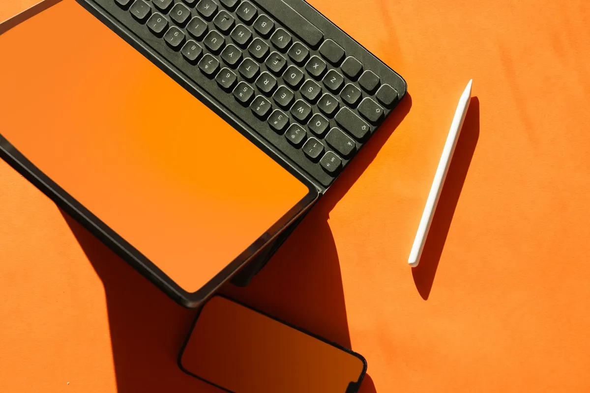 Stylish top-down view of a tablet with keyboard, smartphone, and stylus on a bright orange backdrop.