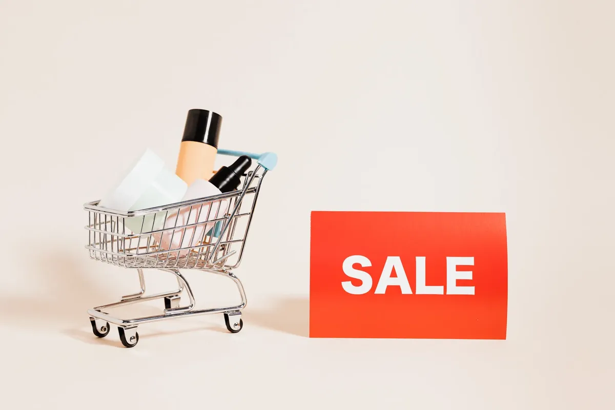A miniature shopping cart filled with products beside a bright sale sign, ideal for retail and marketing concepts.